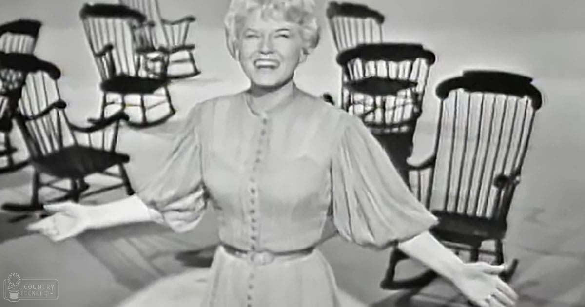 A middle-aged woman wearing a long-sleeved blouse with puffed sleeves in an indoor setting; multiple wooden chairs and light-colored background suggest daytime conditions.