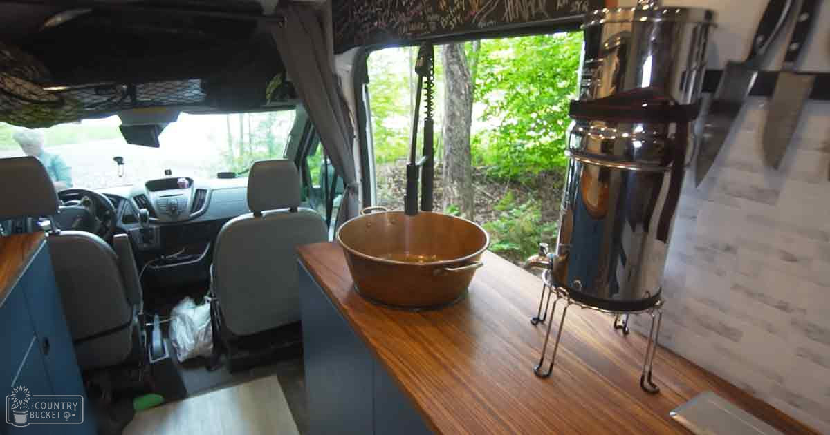 Van interior with wooden countertop and copper pot in a forested outdoor setting during midday.