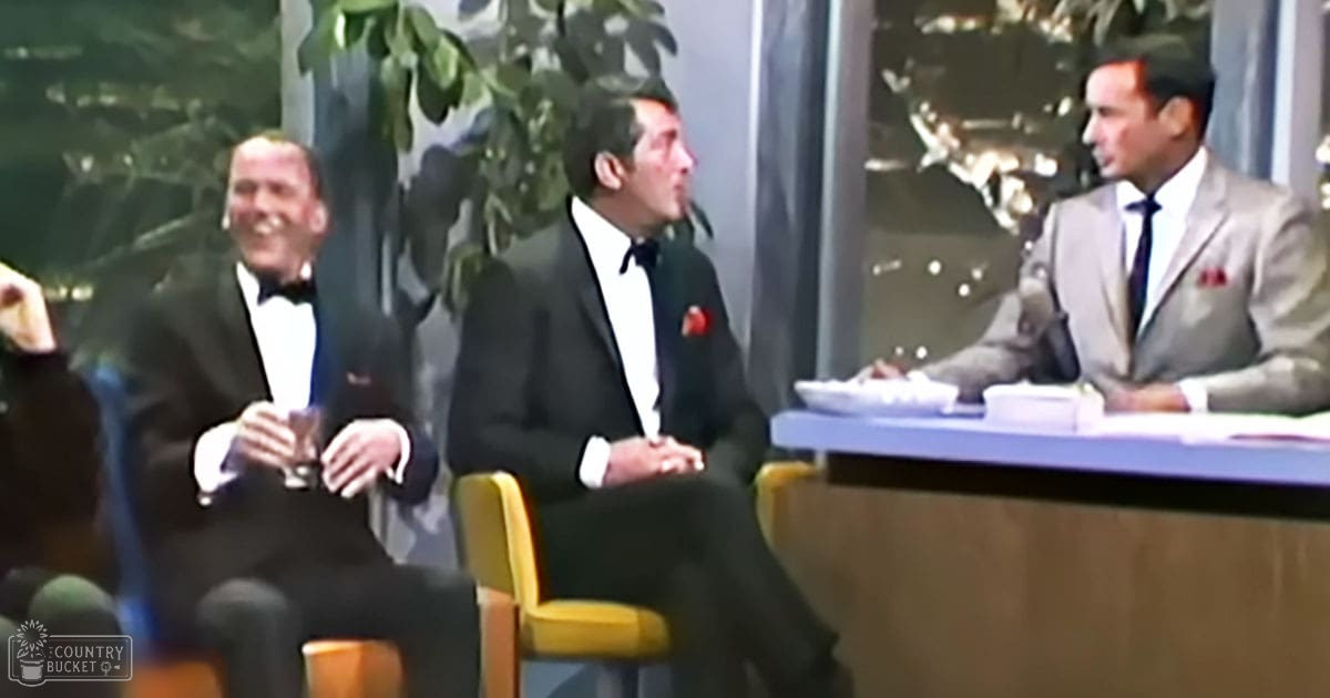 Three men in formal attire seated on a stage, likely during a mid-20th-century talk show, with a host behind them. The setting includes yellow chairs and a wooden desk with greenery in the background.