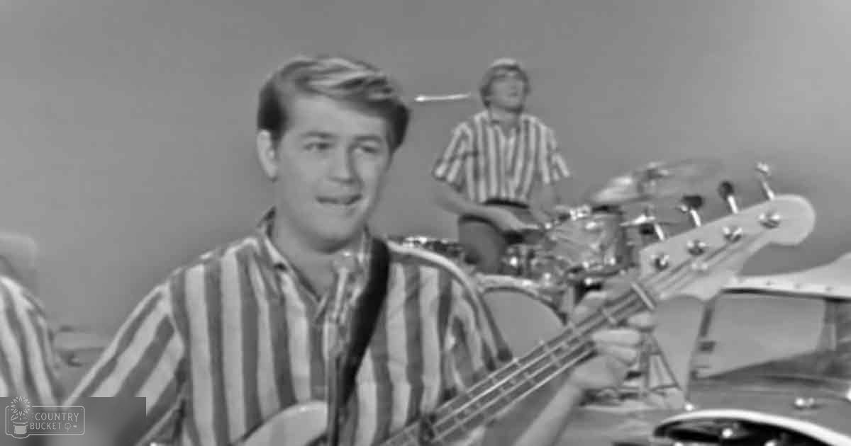 A young adult male playing bass guitar indoors with a microphone setup; another person plays drums in the background.
