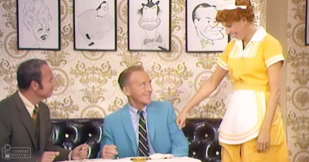 A sitcom or television show scene set in a restaurant, with an adult male in a dark green suit jacket seated left, another adult male in a bright blue blazer seated right, and an adult female in a yellow dress standing right. The setting includes black leather booth seating with framed caricatures of cartoon characters on patterned wallpaper.