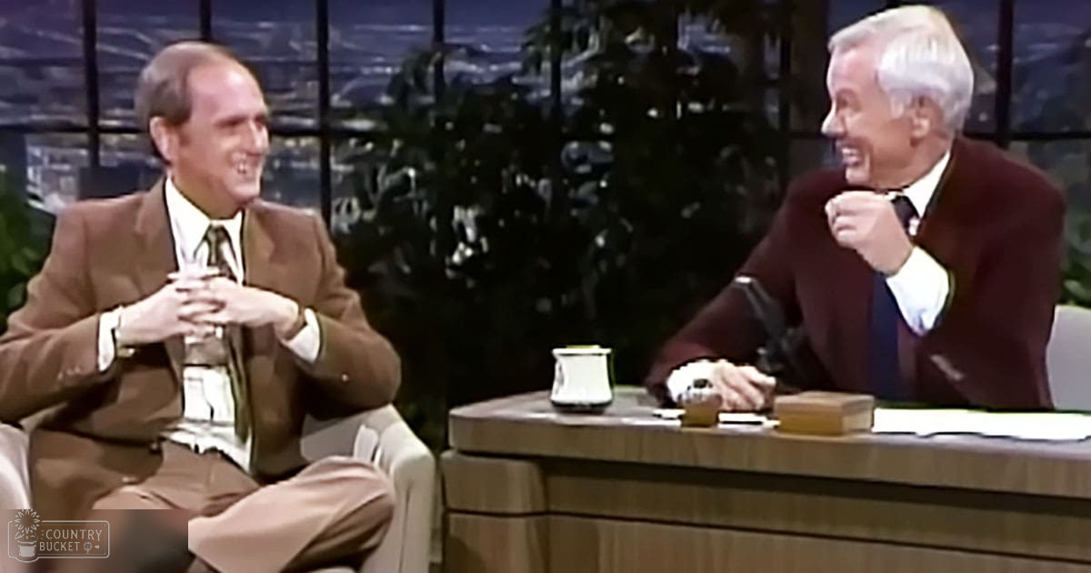 Two adults engaged in conversation in a talk show setting with white armchairs and wooden desk, large windows showing greenery outside, natural light suggests daytime during spring or summer.