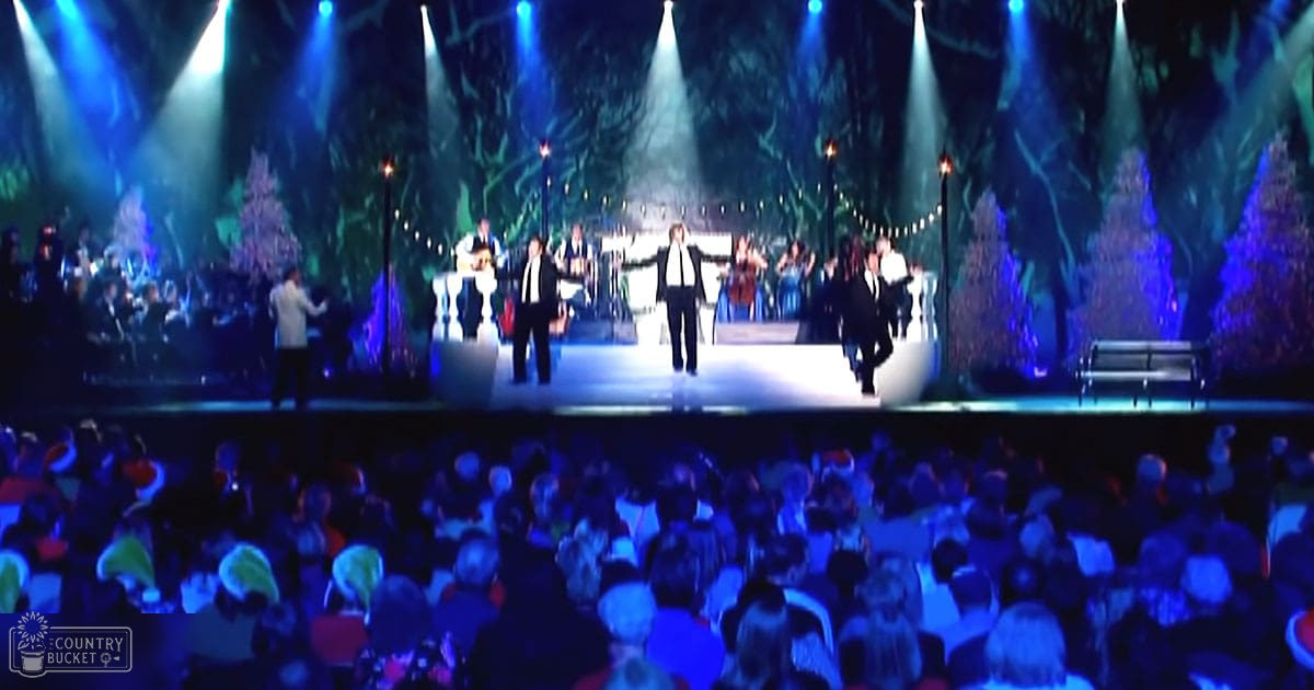 Concert hall stage performance with an orchestra and singers in front of a mixed-age adult audience dressed in formal attire under dramatic stage lighting resembling a forest setting.