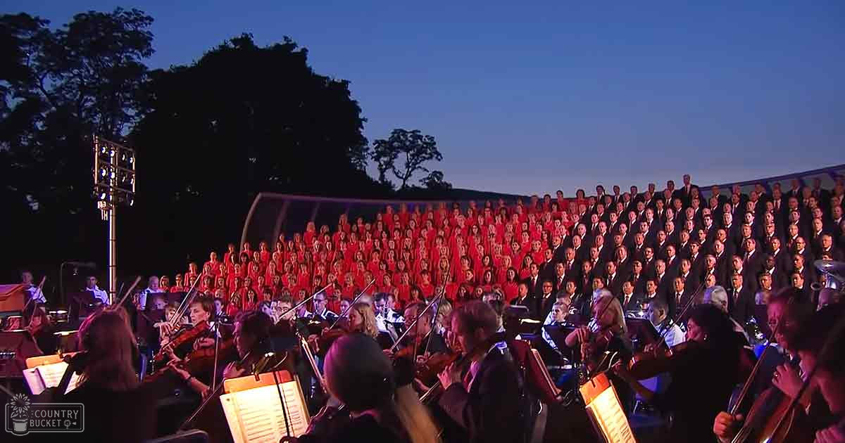 A large choir performing outdoors at dusk with an orchestra in front of them, adults wearing formal attire under a clear sky transitioning from dusk to night.