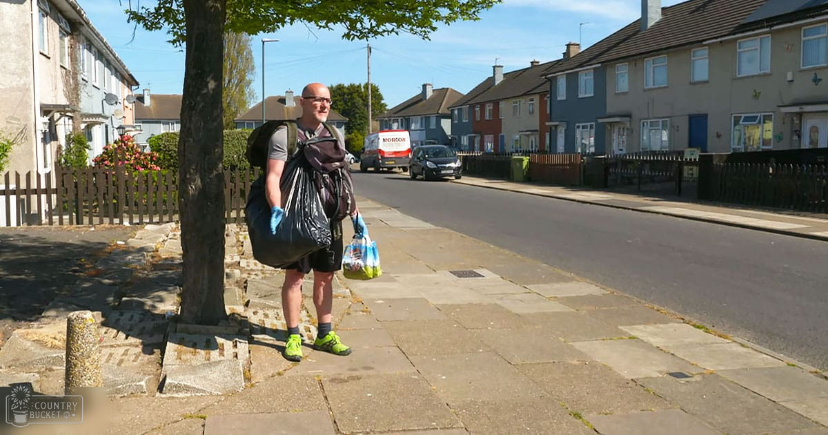 Adult male wearing gray t-shirt, black shorts, green sneakers, blue gloves; carrying trash bags in a residential neighborhood with varying colored houses and parked cars during daytime.