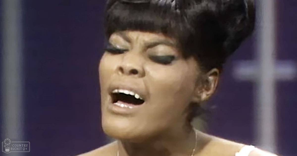 A middle-aged adult female singer in a white strapless top, performing on an indoor stage with purple lighting.