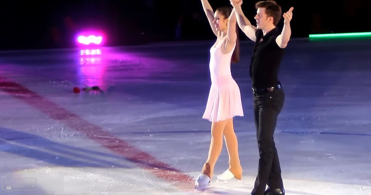 A young adult female figure skater in a light pink dress and white figure skates performs alongside a male counterpart in a black shirt and dark pants, both on an indoor ice rink with colorful stage lighting.