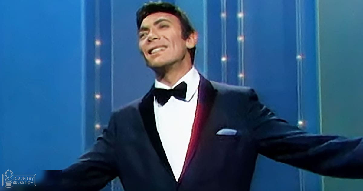 A young adult male in a formal setting, wearing a black tuxedo with white shirt and burgundy vest, standing against blue curtains and string lights.