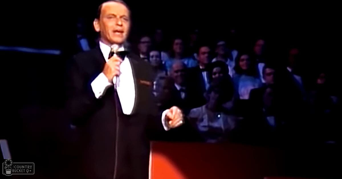 An older adult male performer in a black tuxedo with white lapels, holding a microphone on stage at what seems like a concert or performance event.
