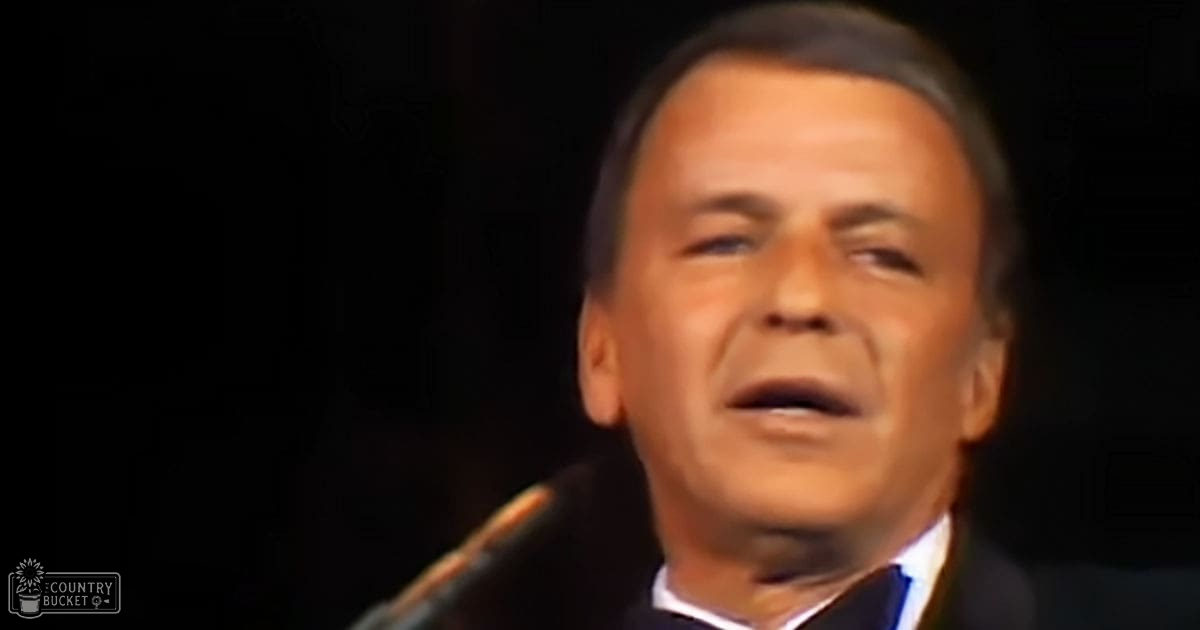 A middle-aged adult male wearing a dark suit jacket and white shirt stands indoors with a microphone near his mouth, suggesting he might be speaking or singing.