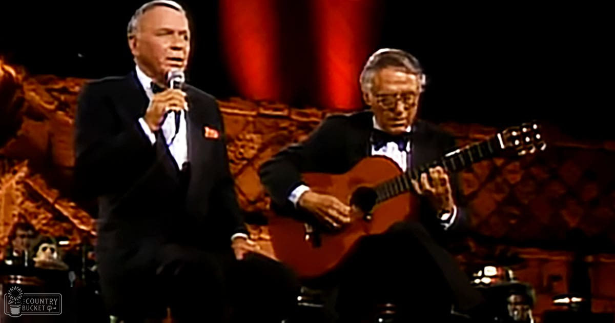 An older adult male singer wearing a dark suit and white shirt, holding a microphone while performing on stage with another musician playing an acoustic guitar in an ornate indoor concert setting.