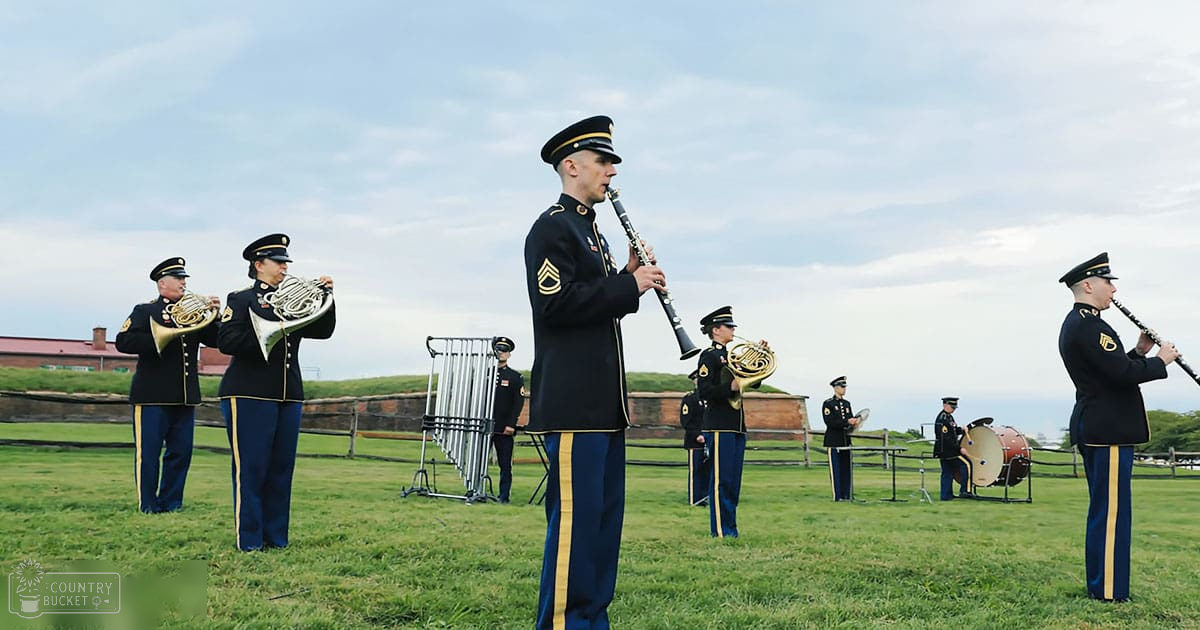 A military band performing outdoors at Fort McHenry with tubular bells and drums, under a partly cloudy sky.