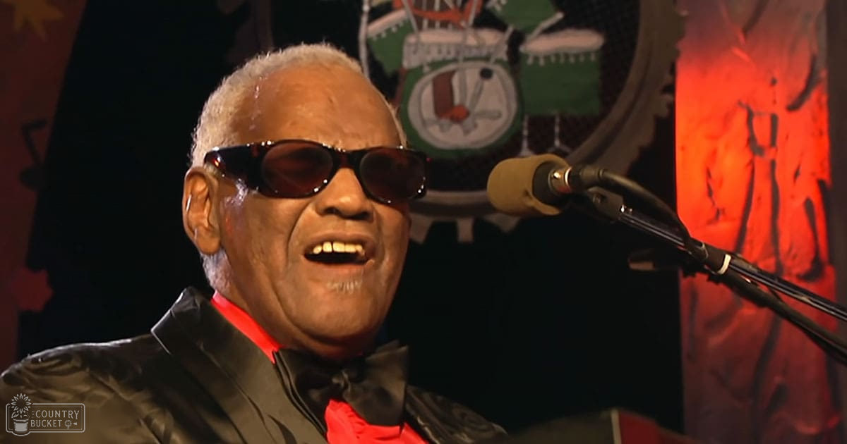 A middle-aged man in a black leather jacket and red shirt stands on stage, wearing a bow tie. The setting includes a decorative background with musical symbols, suggesting a performance or event.