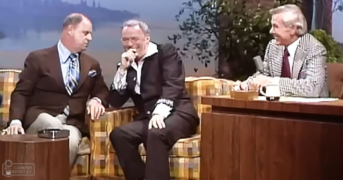 Outdoor talk show set with three men seated in chairs, scenic backdrop of trees and possibly mountains, evening lighting.