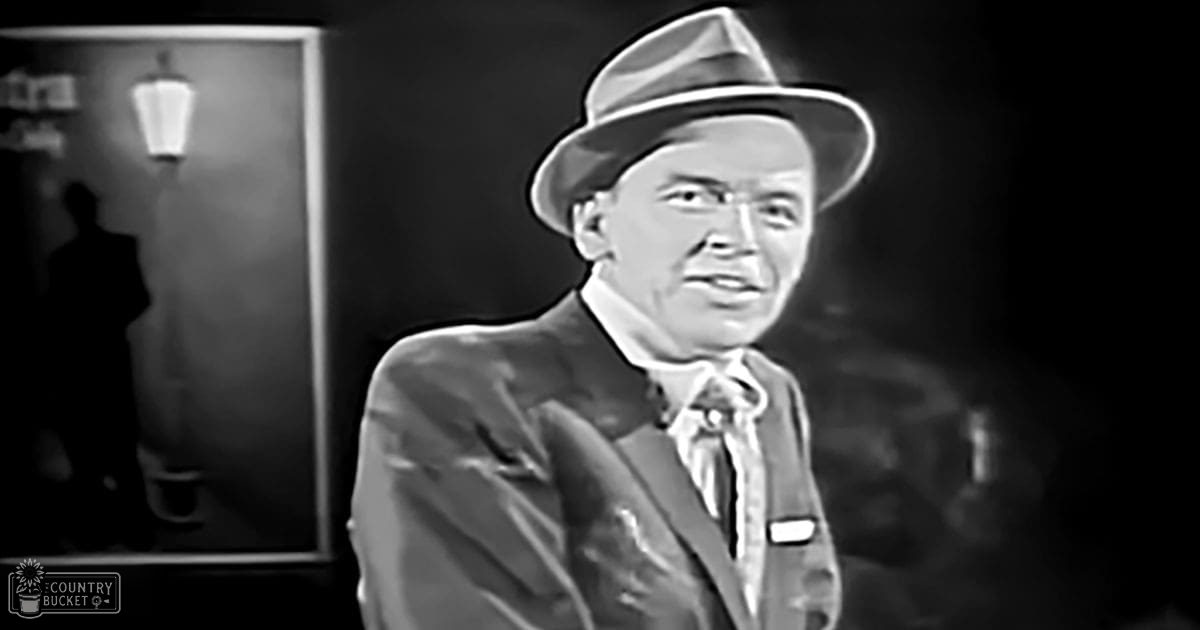 A middle-aged male in a formal dark suit jacket with light-colored shirt and tie, wearing a fedora hat indoors.