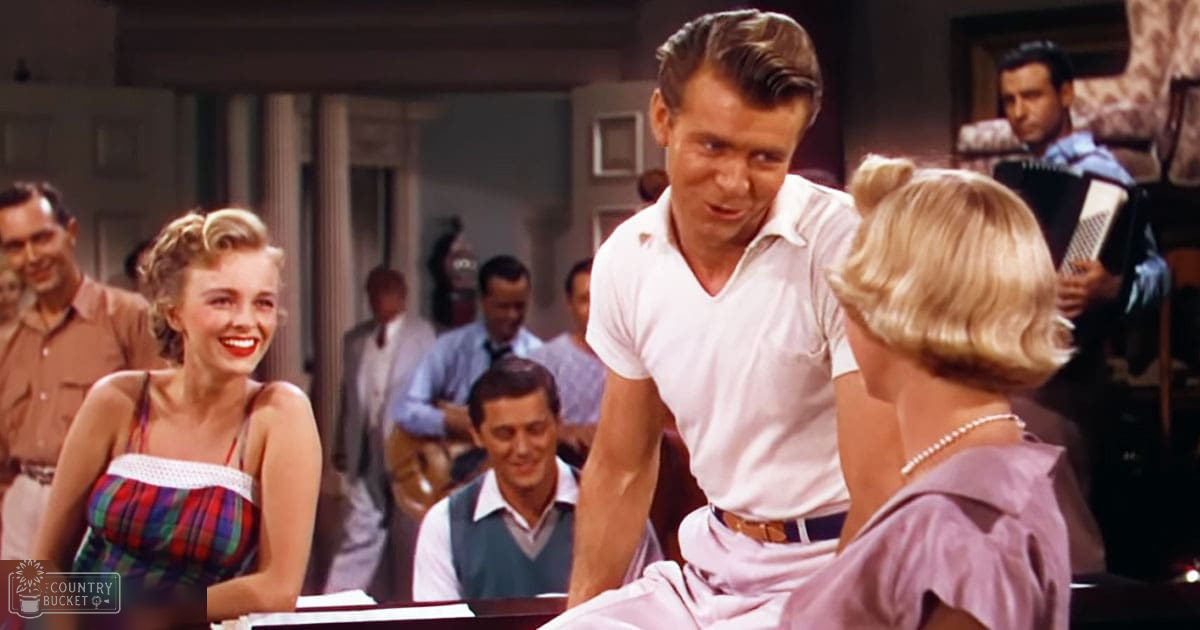 A young adult male in a white short-sleeved shirt and light-colored pants, holding an accordion-like instrument, surrounded by other adults at a bar or social gathering setting with tables and chairs.