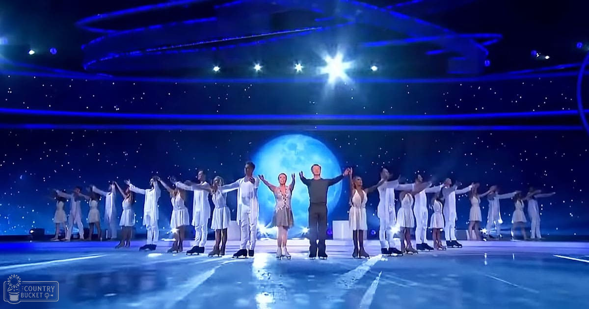 An ice skating performance in an indoor arena with a large moon backdrop, illuminated by starry sky lighting effects. Performers are dressed in white and dark costumes, with synchronized movements on the reflective surface.