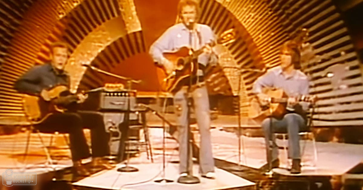 Adult males performing on a stage with microphones, stools, an amplifier, and a pedal steel guitar, set against a sunburst backdrop under artificial lighting in what appears to be a studio or performance venue.