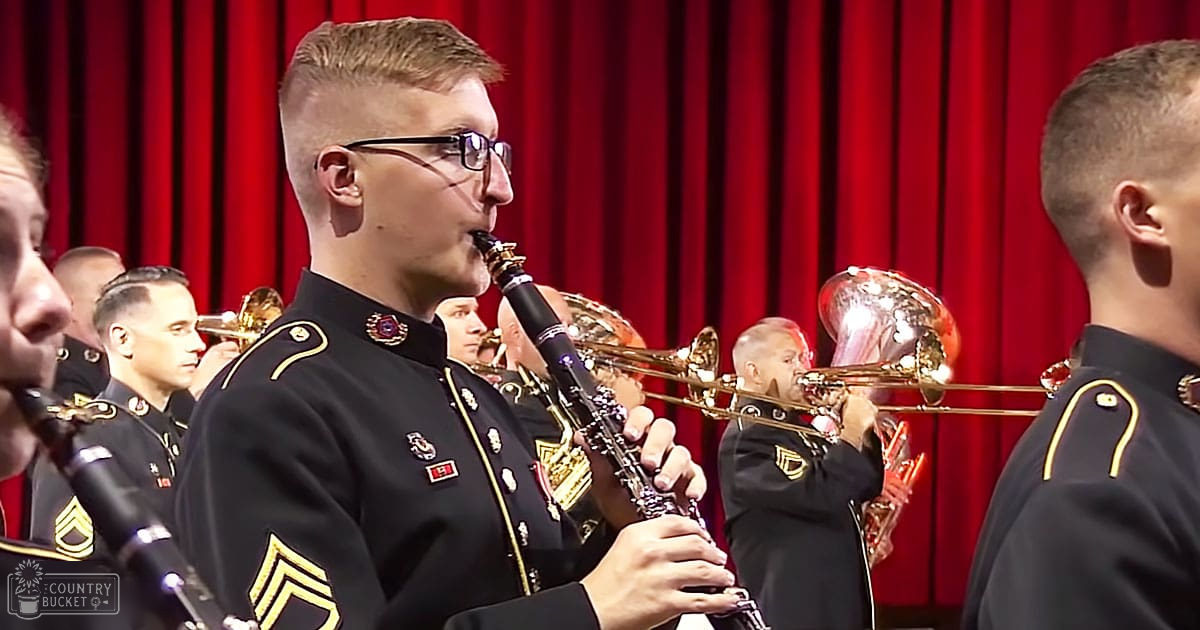 Young adult male playing a clarinet in a military band setting with red curtains and bright indoor lighting.