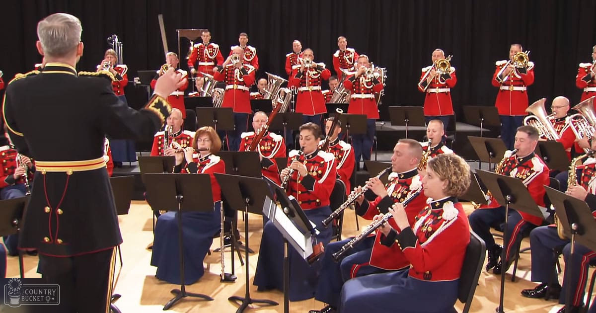 A military band performing in a concert hall with seated musicians wearing red uniforms and gold accents, under warm stage lighting.