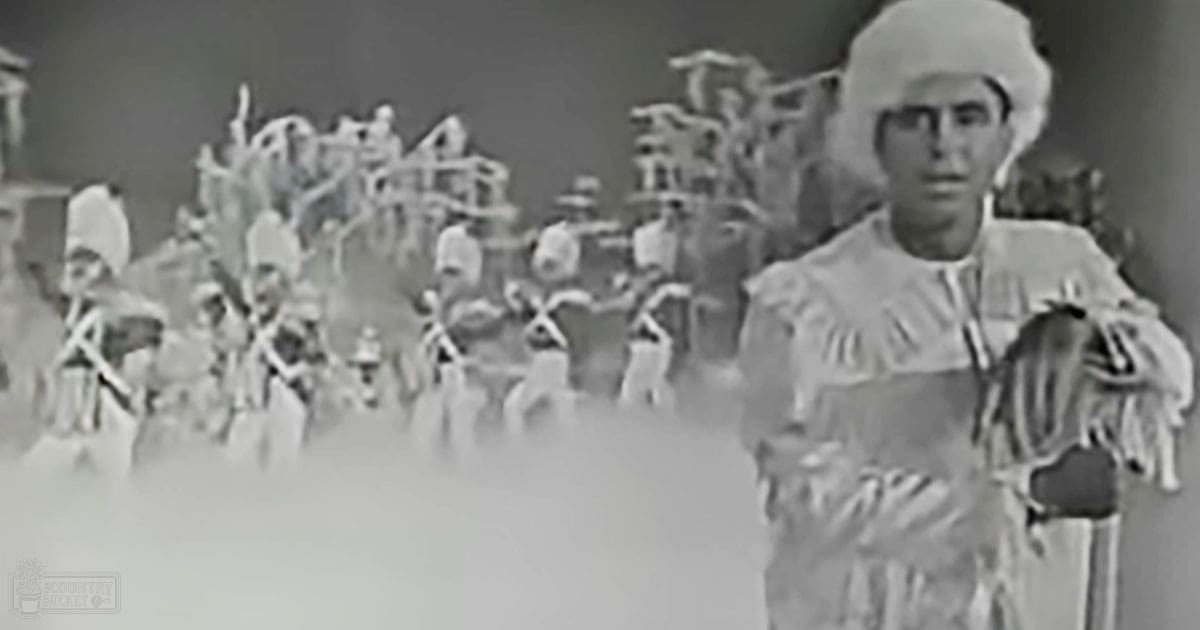 A young adult male wearing a white turban-style head covering and ruffled sleeves in an indoor setting; blurred background suggests studio or stage.