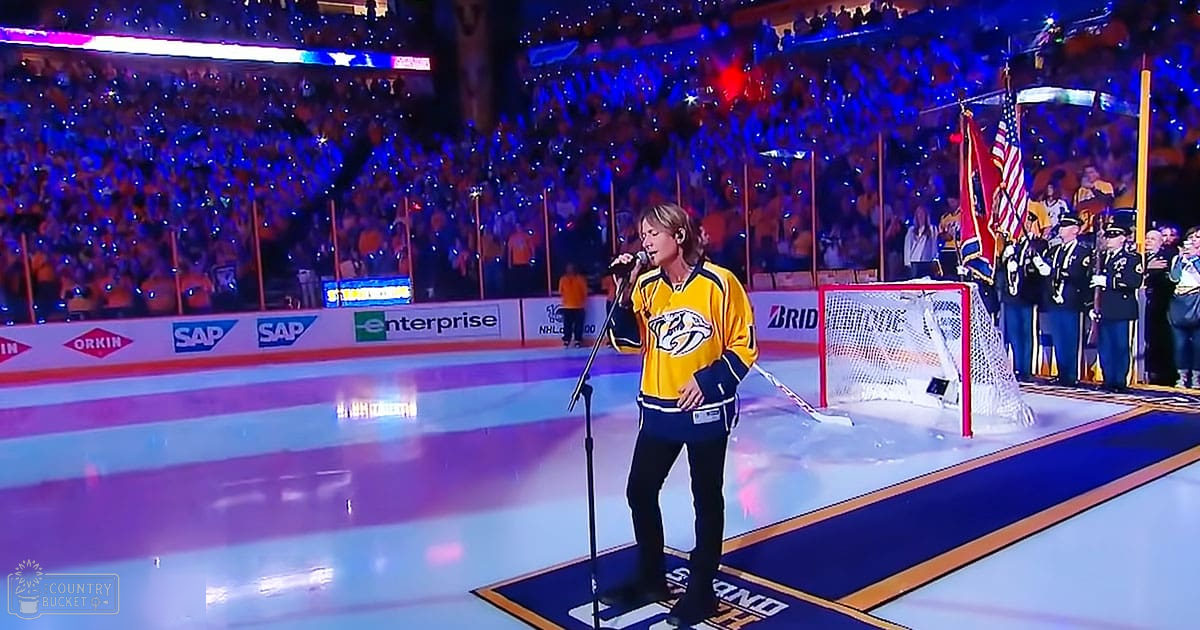 An adult male singer performing at an indoor ice hockey arena with spectators holding up lights during a pre-game ceremony.