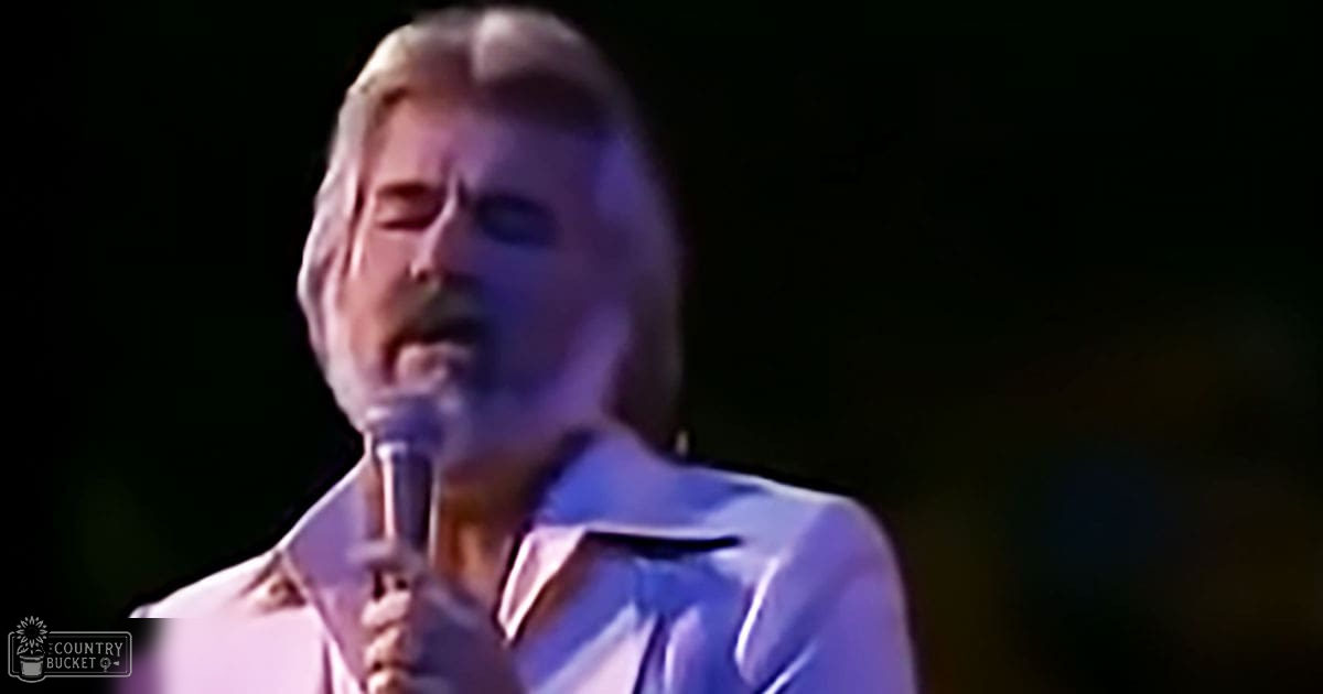 Middle-aged man singing into a microphone in an indoor performance setting under dim stage lights.