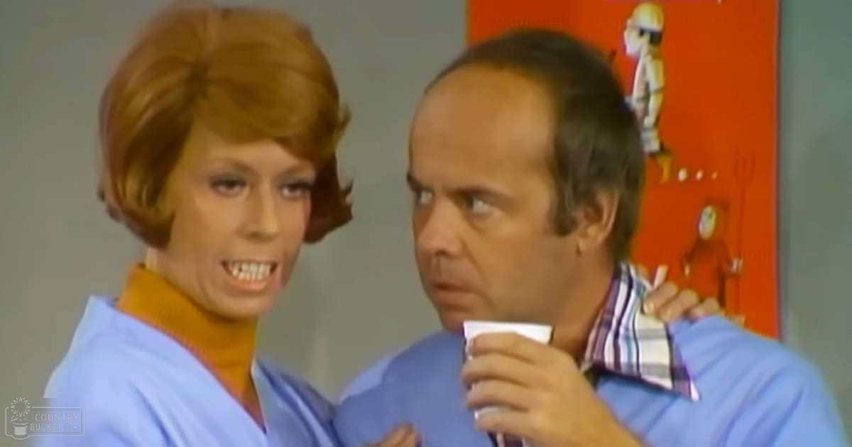 An indoor television or movie set with an adult female wearing a light blue top and an adult male in a rolled-up sleeve plaid shirt, surrounded by decorative items on the wall.