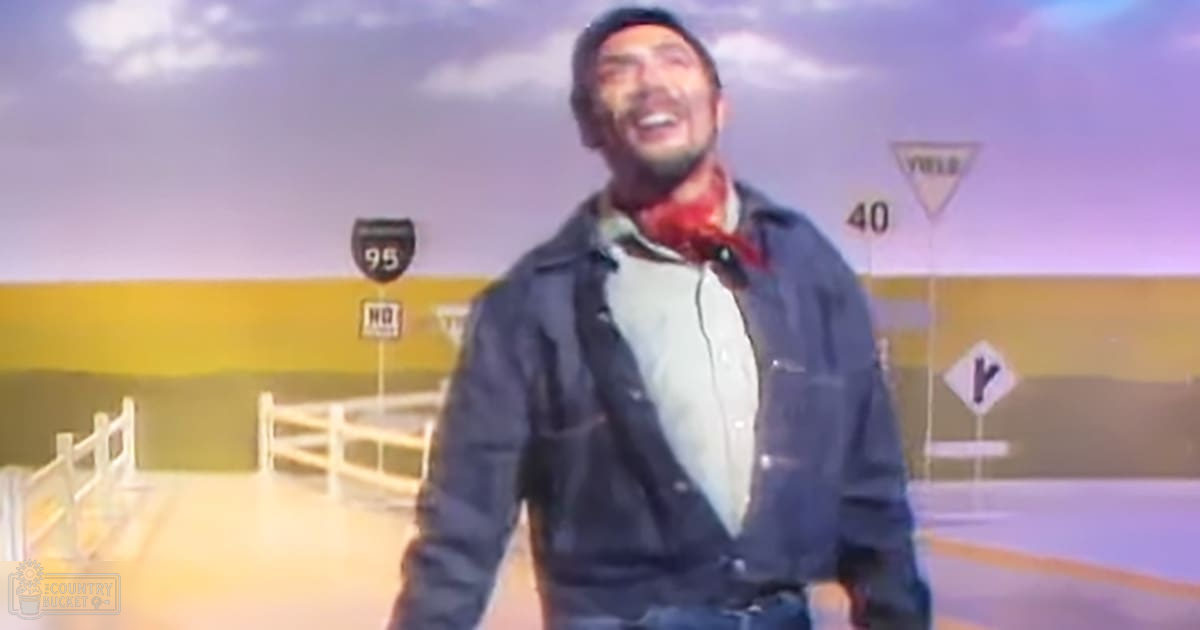 A young adult male stands outdoors in a rural highway scene painted backdrop, wearing a dark blue denim jacket with light-colored buttons and pockets, a white shirt underneath, and a red bandana around his neck. The setting includes road signs such as '95,' 'NO,' 'YIELD,' and '40,' under soft lighting suggesting early morning or late evening.
