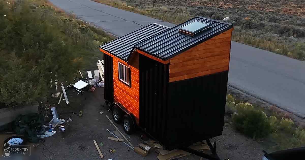 A tiny house with a black roof on a paved road surrounded by construction materials and vegetation at dusk.