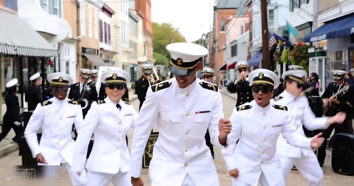 Young adults in white military-style uniforms dancing outdoors during a parade or public event with historical architecture, flags, and urban surroundings.