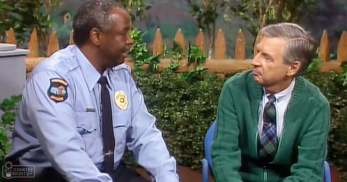 Adult male police officer in mid-20th-century light blue uniform outdoors with older adult man in green cardigan, white collared shirt, and plaid tie. Setting features a wooden picket fence and lush greenery under natural daylight.