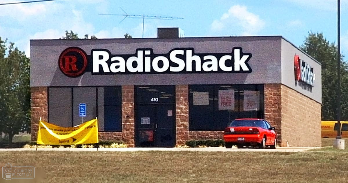 A RadioShack storefront with a 'Sprint' banner, parked red car and yellow DHL delivery van on an urban street lined by trees under clear sky.