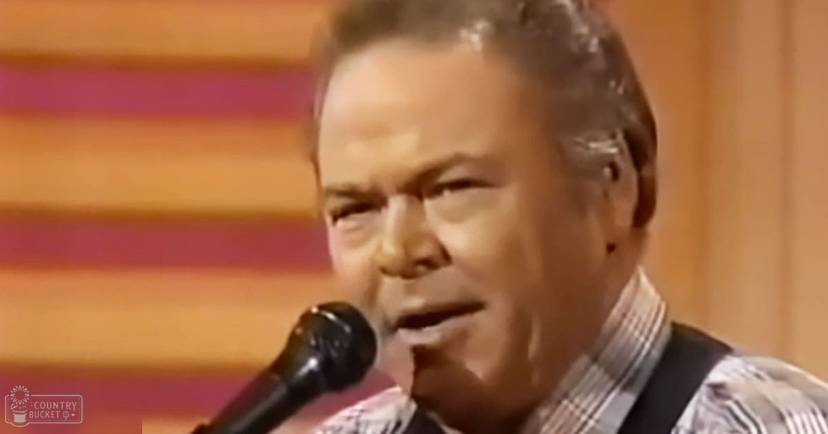 A middle-aged adult male wearing a plaid shirt in an indoor setting with warm lighting, possibly on a stage.