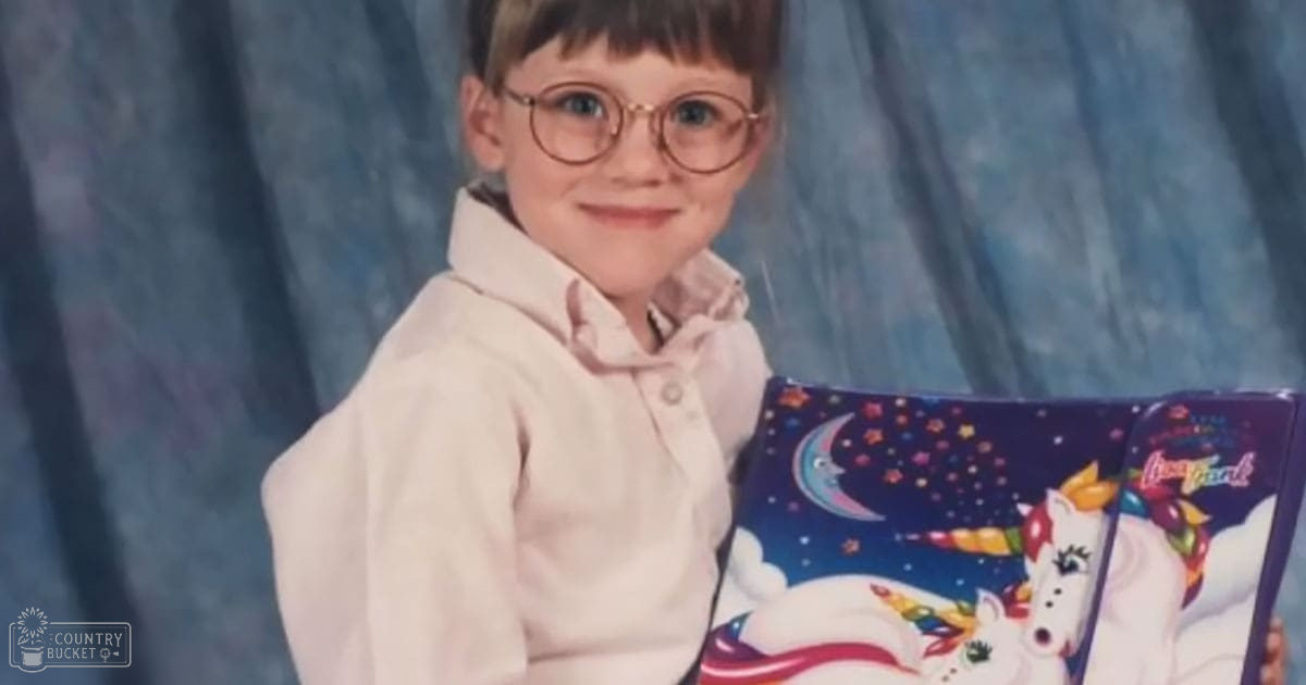 A young child wearing a light-colored button-up shirt, holding an open book with colorful unicorn and star illustrations on a plain blue curtain backdrop.
