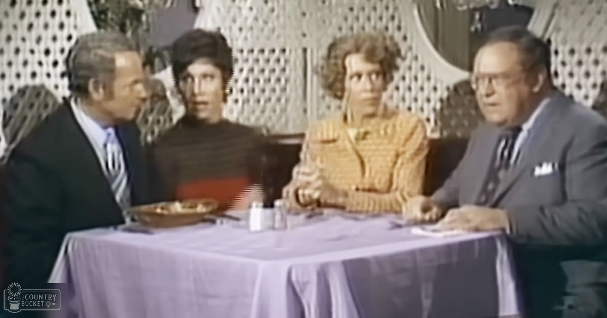 A group of adults engaged in conversation around a dining table, set against patterned wallpaper or curtains, with light purple tablecloth and food items on the table.