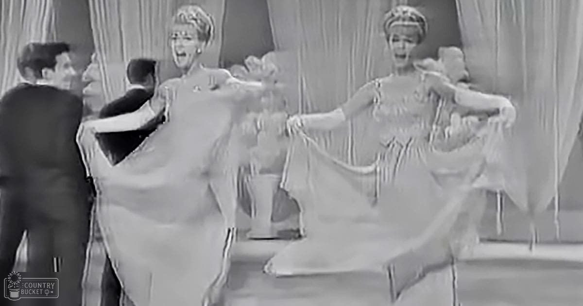 A dance performance scene with two adult females in light-colored dresses from an earlier era performing on a stage set against curtains under bright studio lighting.