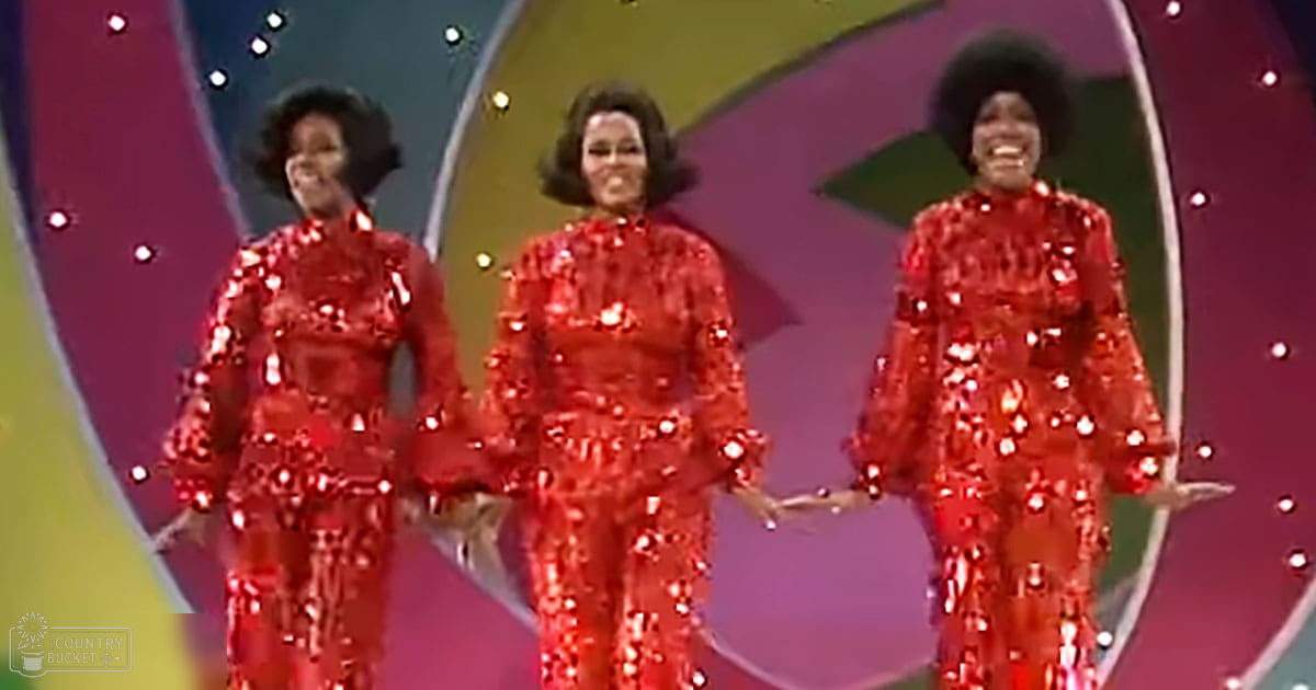 A group performance scene with three adult female performers in matching red sequined outfits on a brightly lit stage with vibrant colors, indicative of a 1970s or early 1980s performance setting.