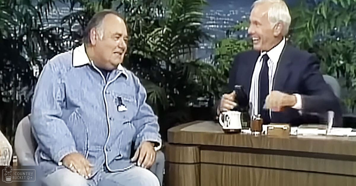 A talk show setting with a middle-aged male wearing a light blue denim jumpsuit and an older adult male in a dark suit jacket, both seated at a wooden desk surrounded by artificial greenery indoors under bright lighting.