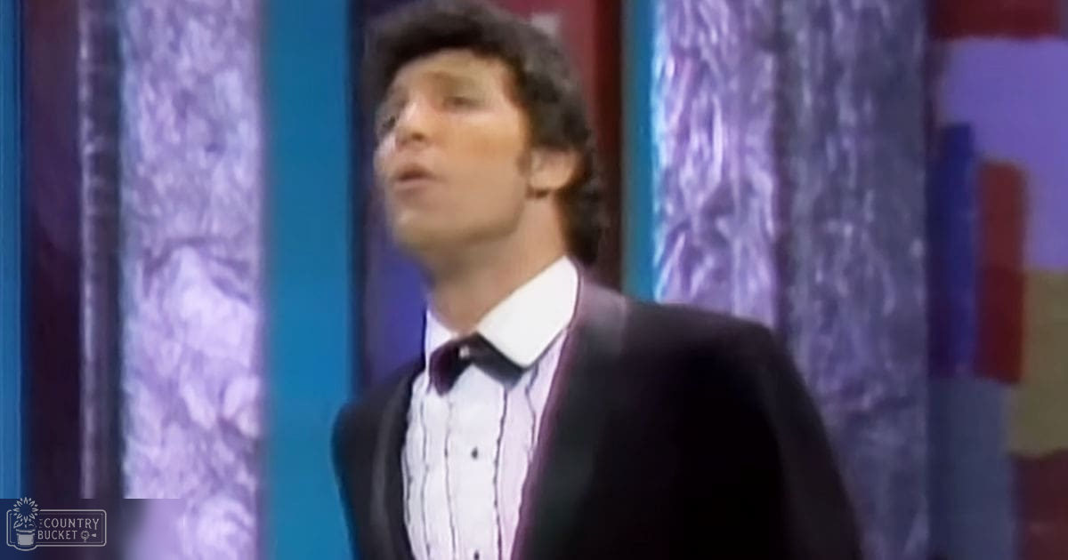 Young adult male wearing a dark suit jacket, white dress shirt with vertical stripes and maroon bow tie in an indoor setting with patterned curtains.