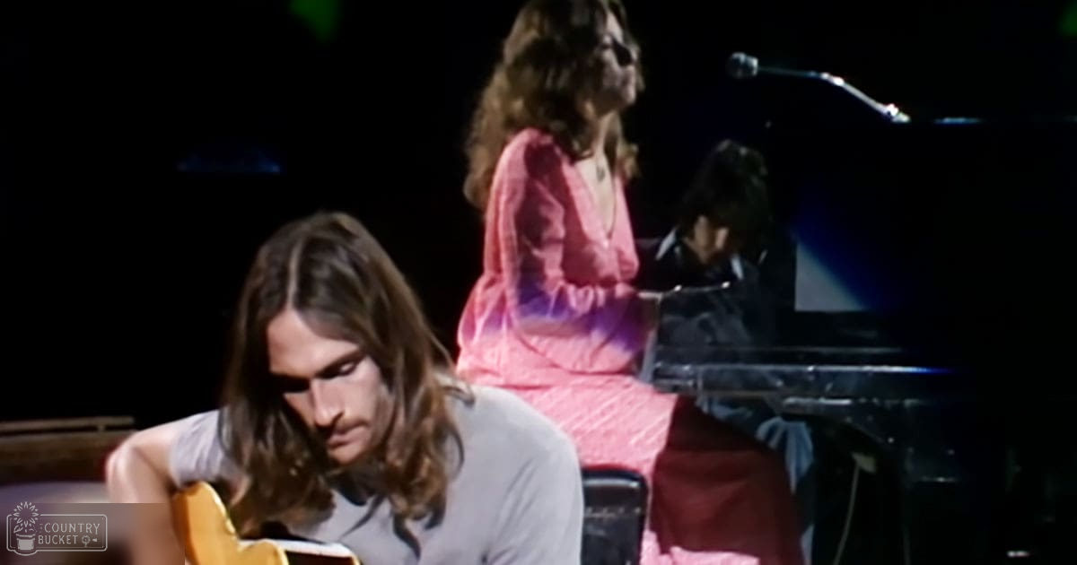 A young adult male musician plays an acoustic guitar while another adult female singer performs into a microphone at what appears to be a concert venue.