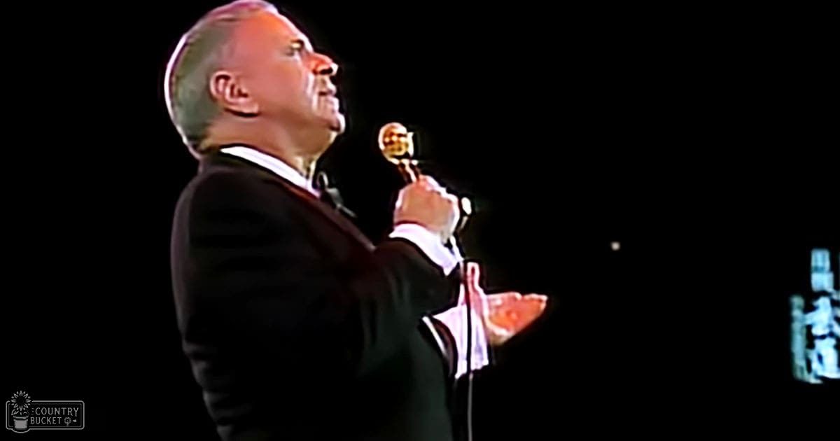 A middle-aged man in a formal dark suit and white shirt holds a golden trophy, possibly at an indoor event or performance with artificial lighting.