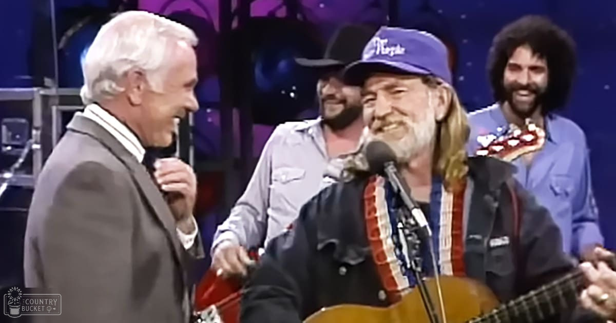 Musical performance scene on a stage with dark curtains and lighting; older adult male musicians in formal attire holding microphones, denim shirts, and acoustic guitars.