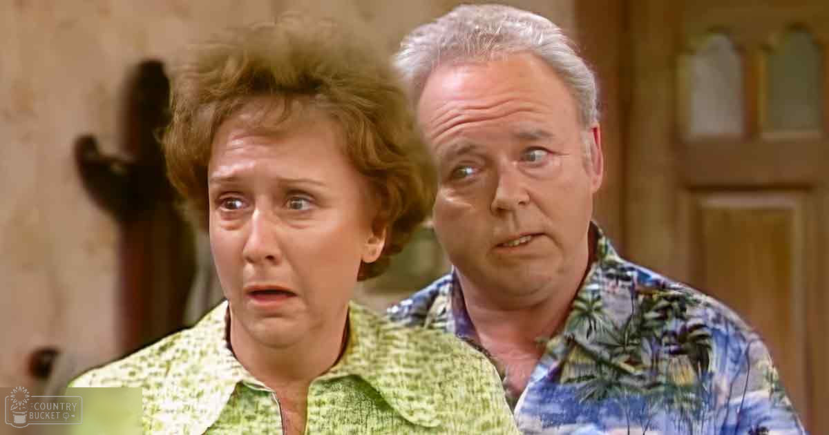 A middle-aged woman in a green patterned shirt stands indoors, accompanied by a middle-aged man wearing a blue palm tree-patterned shirt. The setting appears to be an older home interior with wooden doors and textured walls.