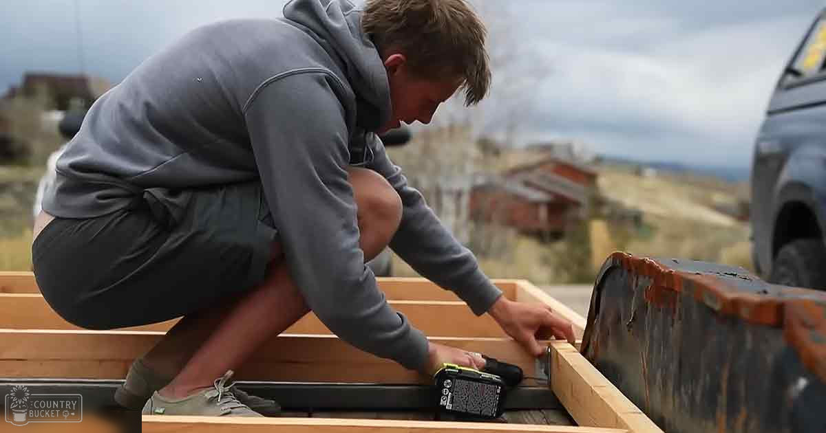 A young adult male works outdoors, focusing on a wooden structure with a black power drill beside him under an overcast sky.