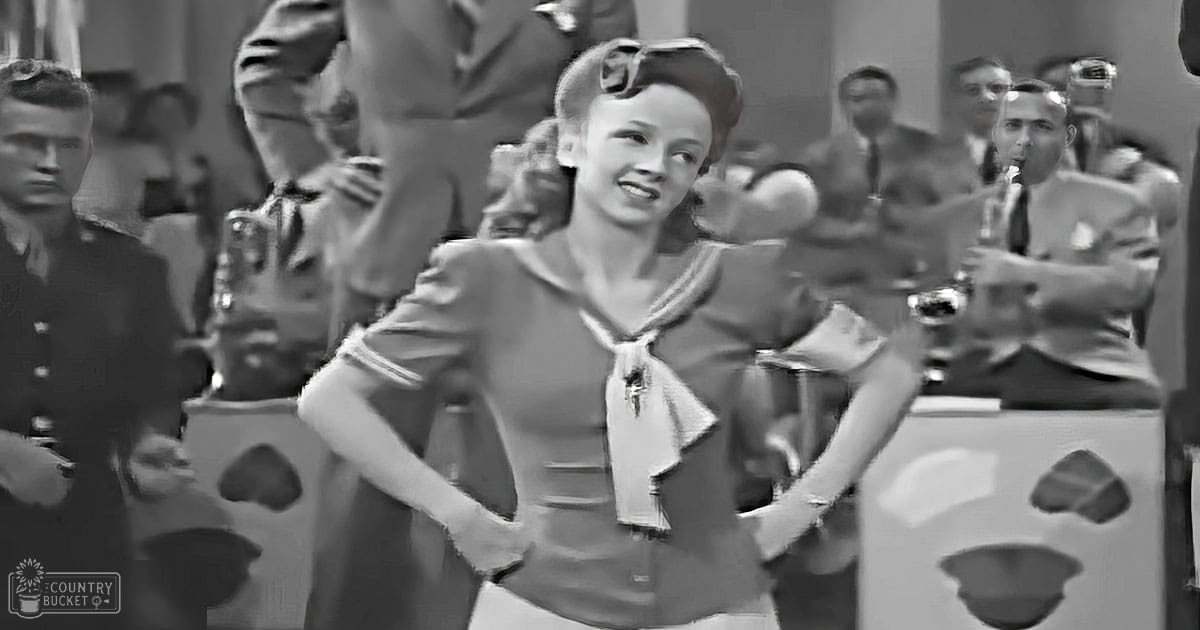 A World War II-era dance scene with a young adult female in a sailor-style blouse and scarf, surrounded by other individuals in military and formal attire within an indoor performance setting.