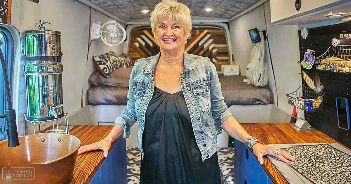 Middle-aged adult female wearing a distressed denim jacket over a black sleeveless top, seated inside an RV interior with visible furniture and home decor elements.