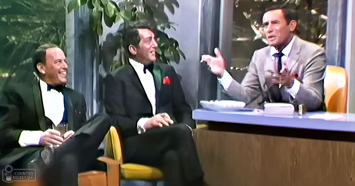 Three adult males seated at a table in an indoor talk show or interview setting with wooden chairs, dark curtains, and decorative plants.
