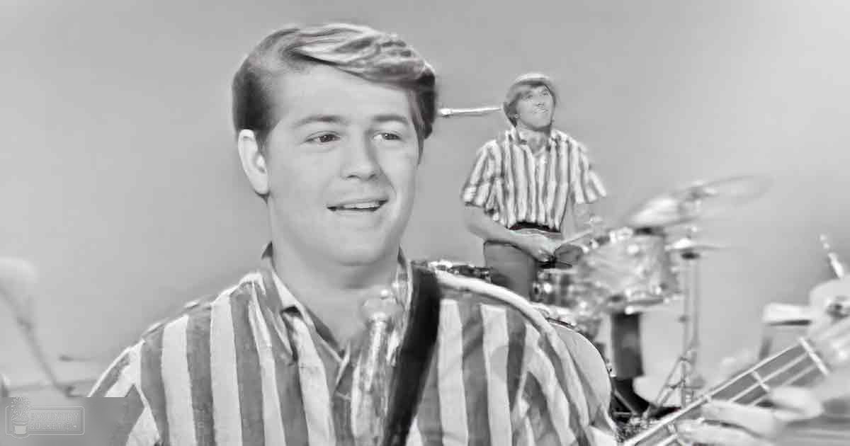 A young adult male in a striped shirt sits indoors with a background person playing drums; simple backdrop suggests performance setting.