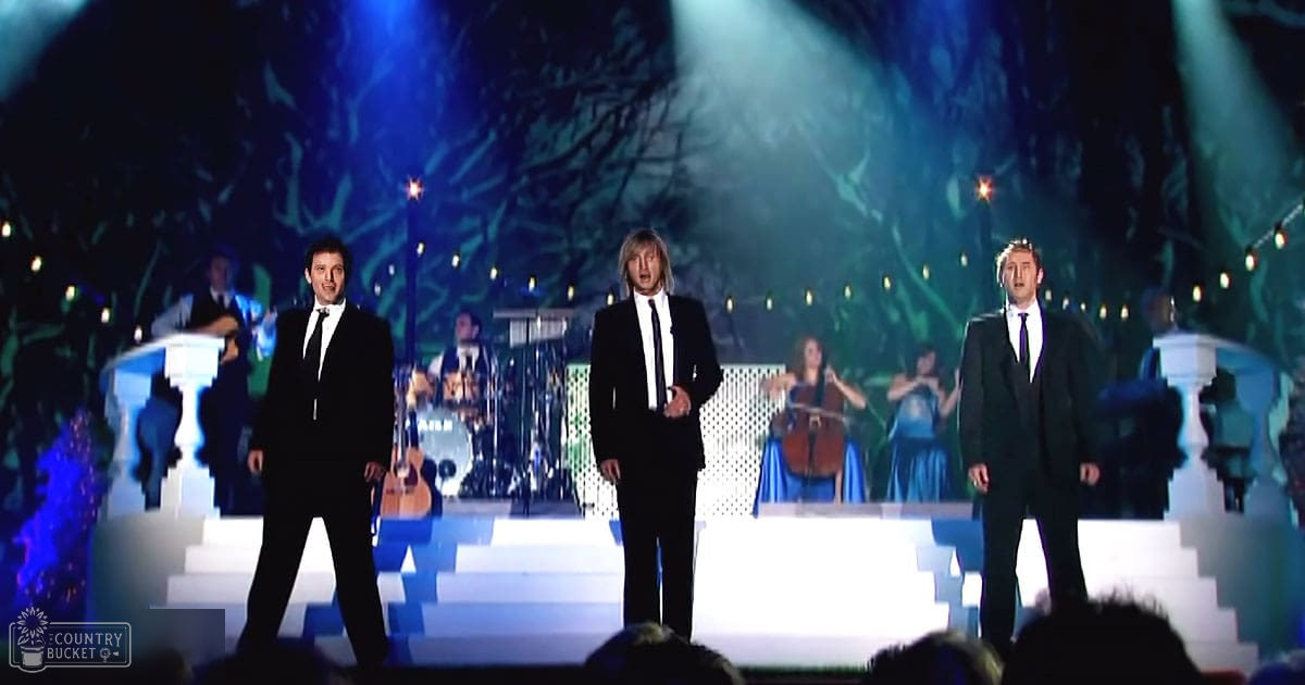 A performance scene at an indoor venue with adult male performers in black suits, stage lighting, musical instruments, and decorative elements creating a dramatic effect.